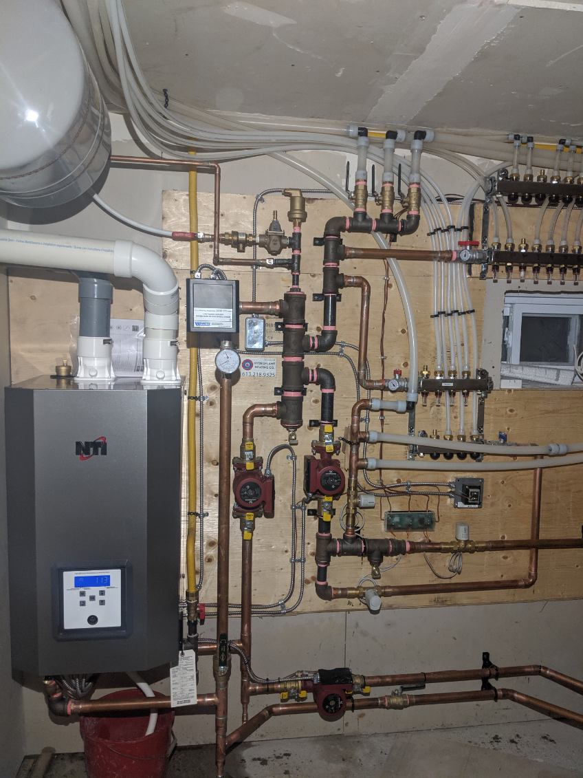 A wall-hung boiler with copper piping, circulator pumps and in-floor heating manifolds on a plywood wall