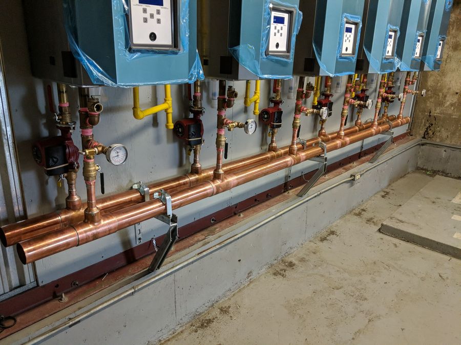 A row of wall-mounted boilers above a copper manifold with circulator pumps and gas piping in a plant room