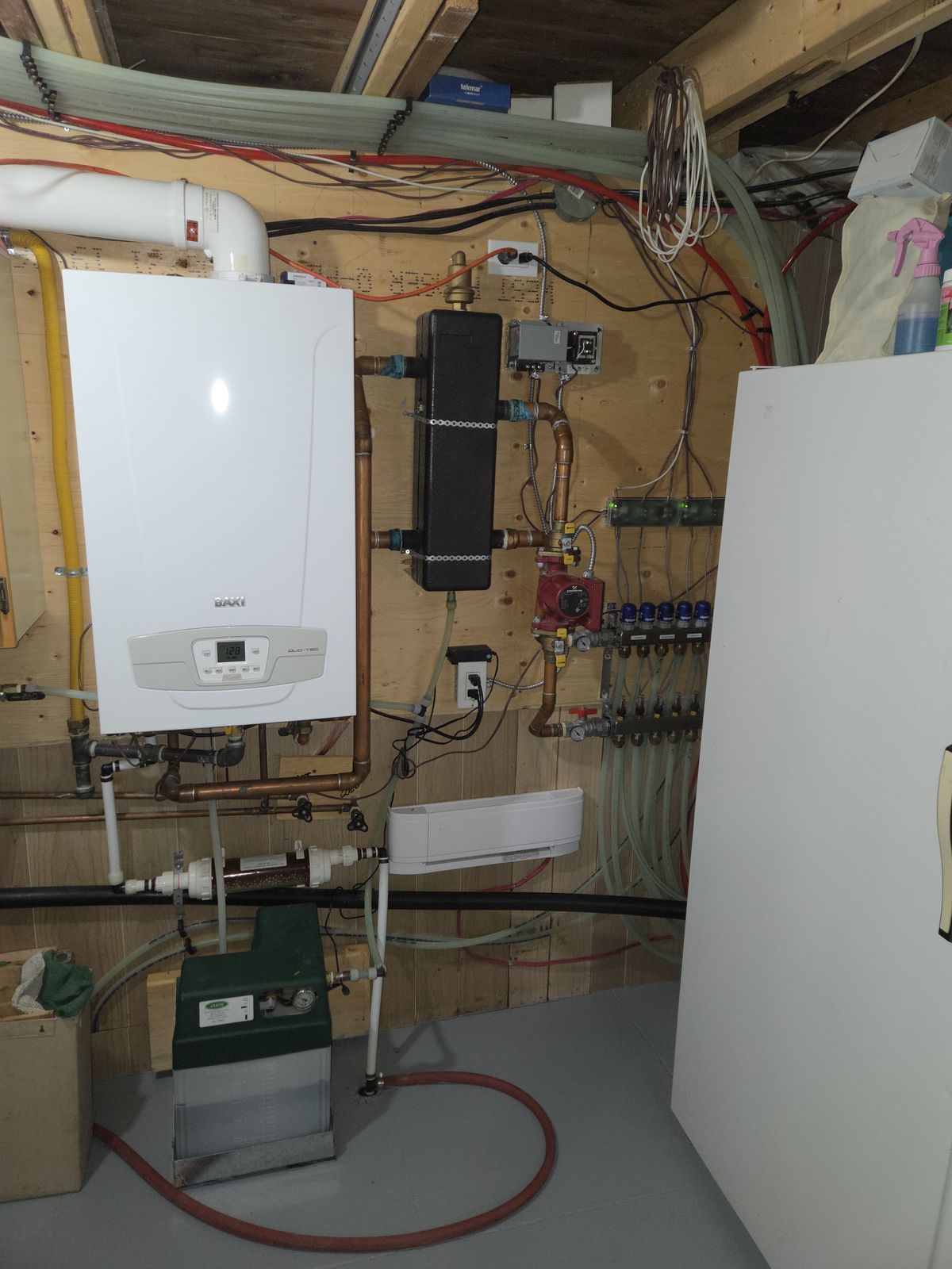 A white wall-hung boiler beside a glycol in-floor heating manifold in a basement mechanical room