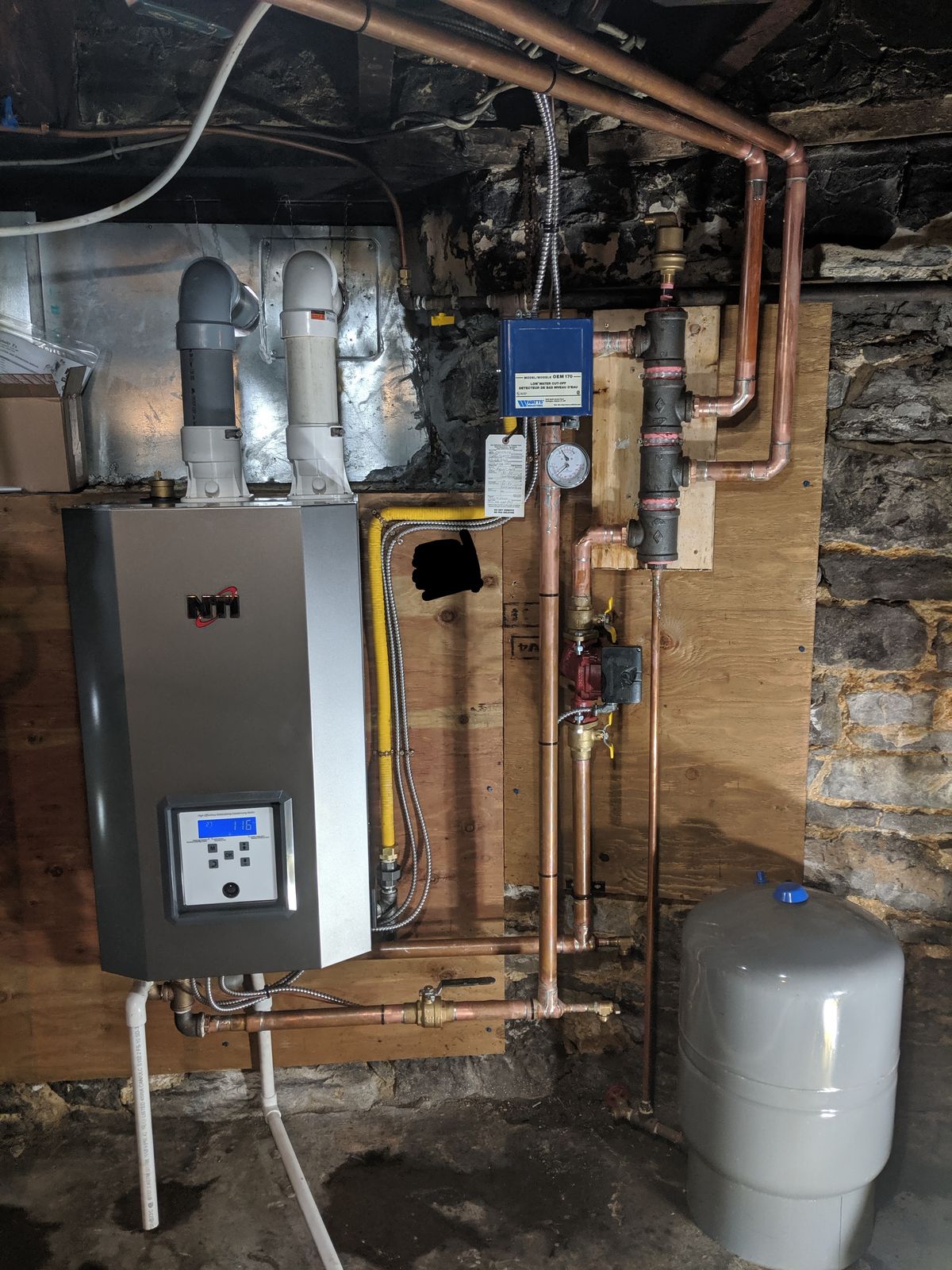 A wall-hung boiler with an expansion tank and copper piping in a stone basement