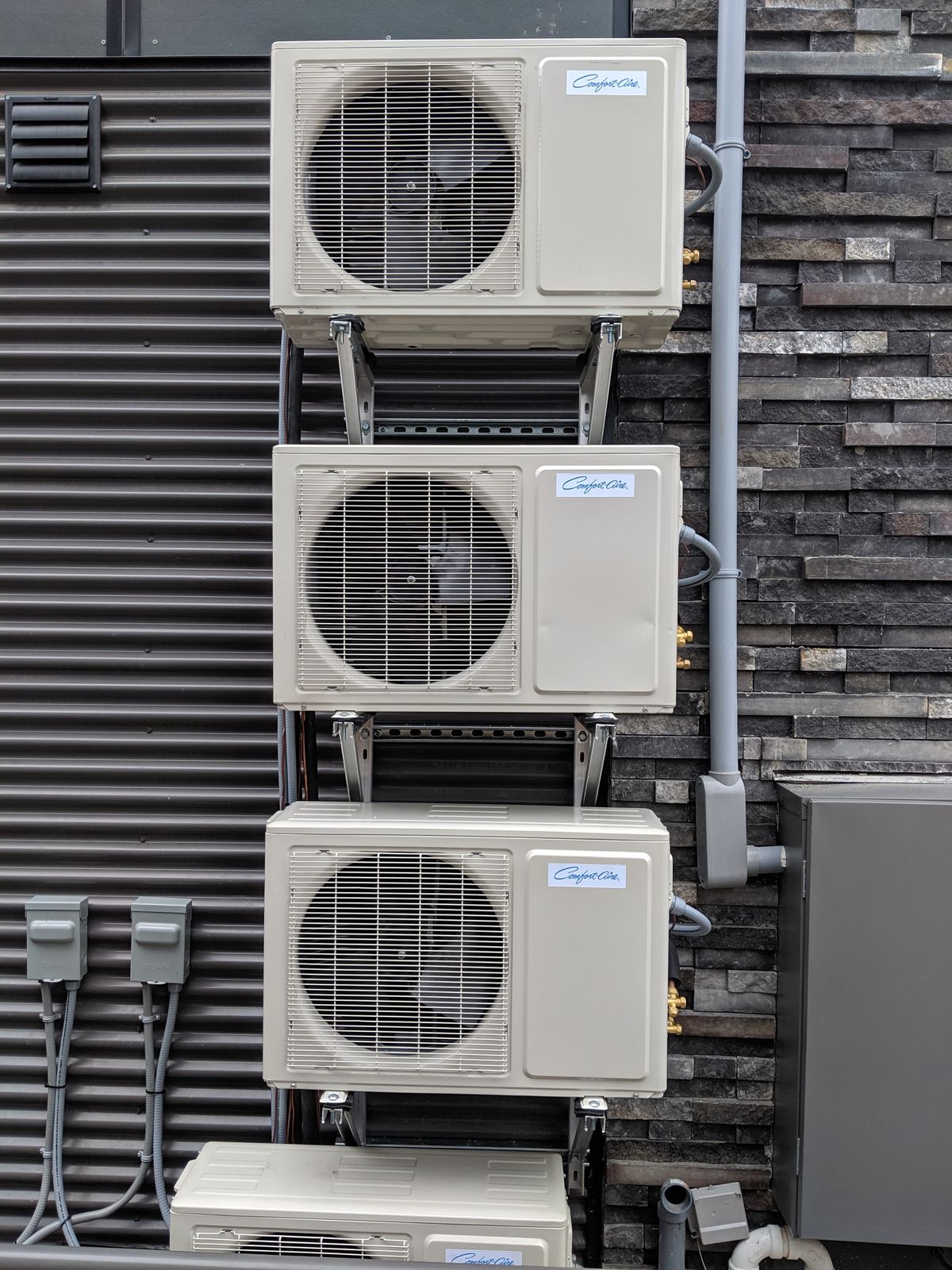 Three ductless outdoor units stacked on wall brackets against a stone and steel-clad wall