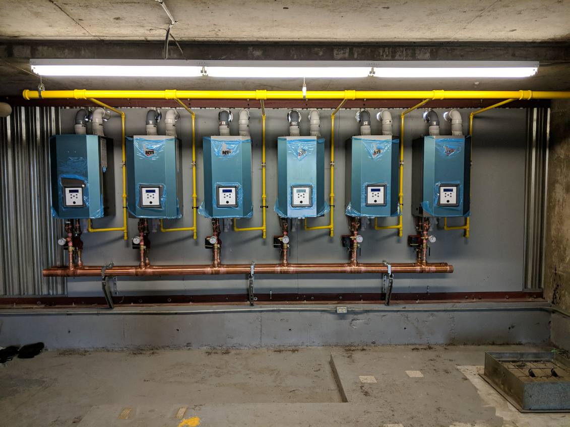 Six wall-hung boilers in a row with yellow gas piping, venting and a copper header in a parking garage mechanical room