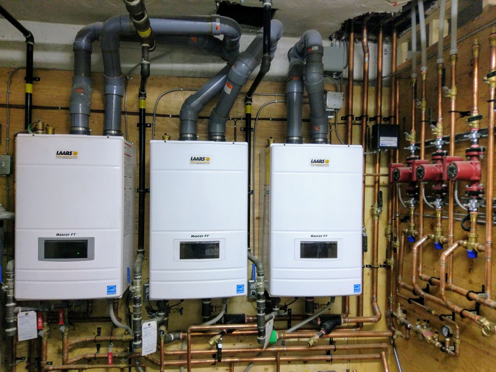 Three white wall-hung combi boilers with venting and copper piping on a plywood wall